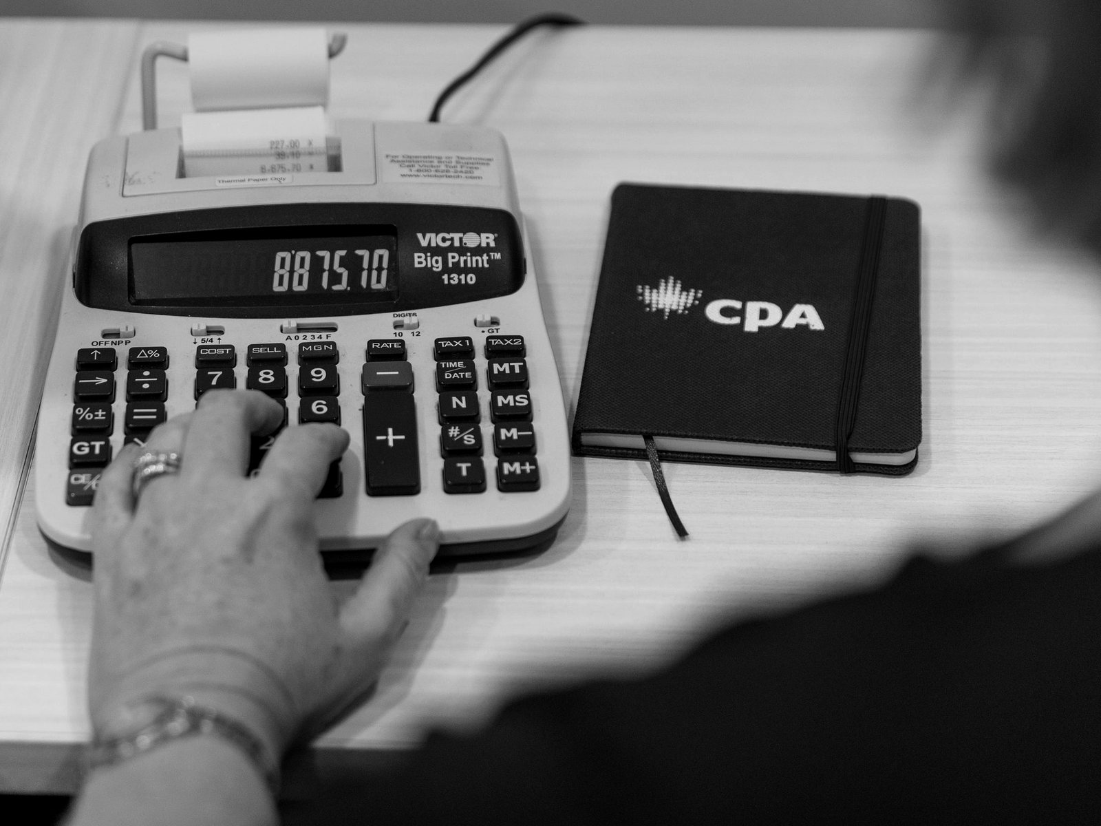 A hand working a printing adding machine on a desk, a notebook beside it.
