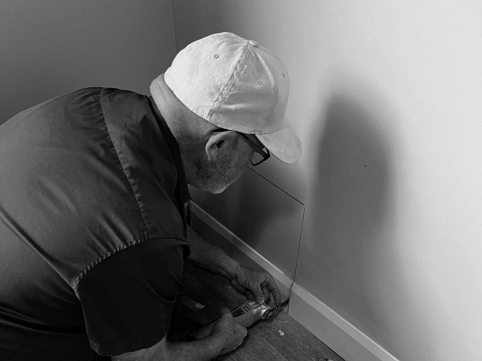 A maintenance technician in a cap kneeling to fix a baseboard in a unit.