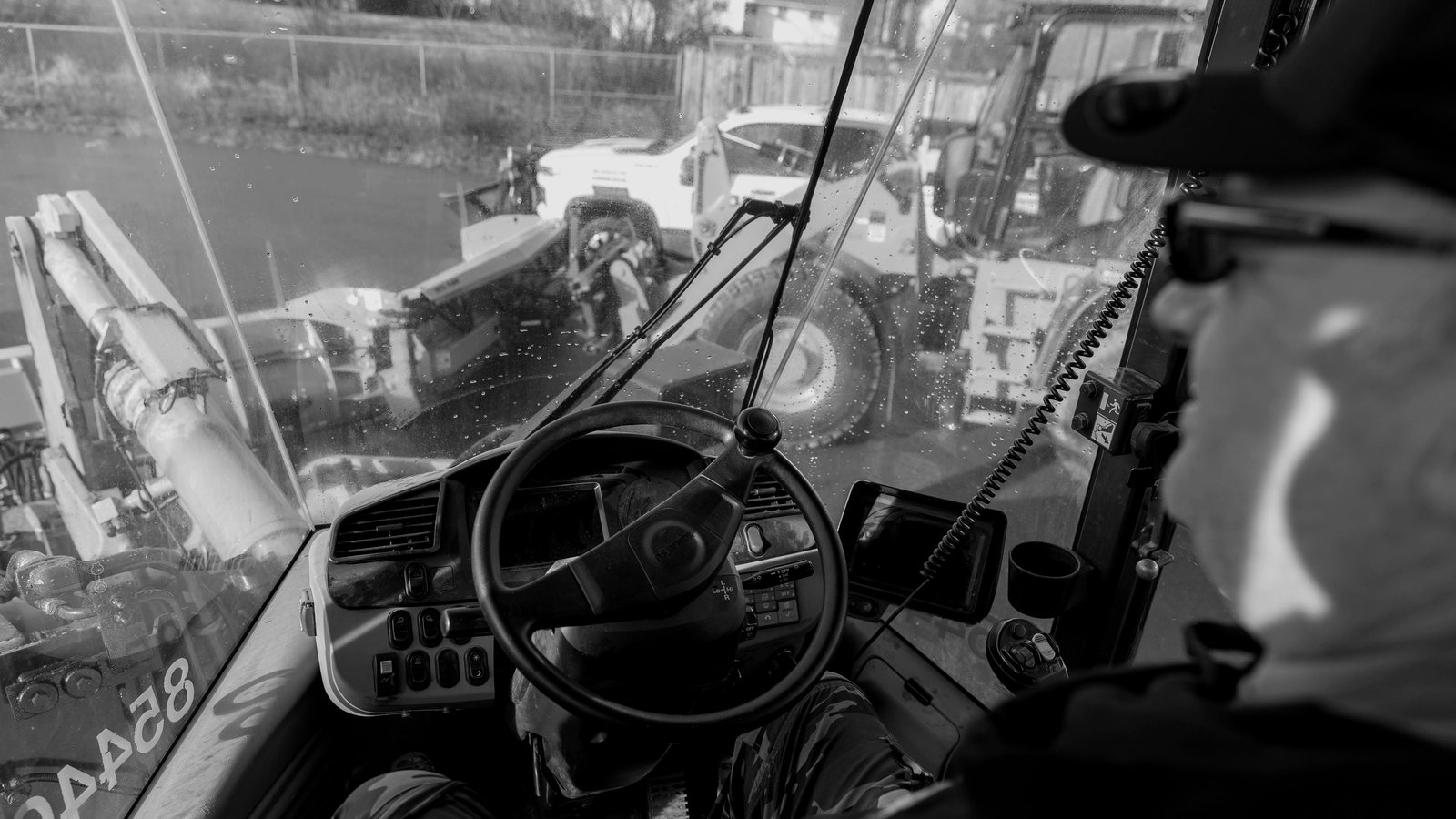 The yard seen over an operator’s shoulder from the seat of a loader: the steering wheel, and more loaders through the windshield.