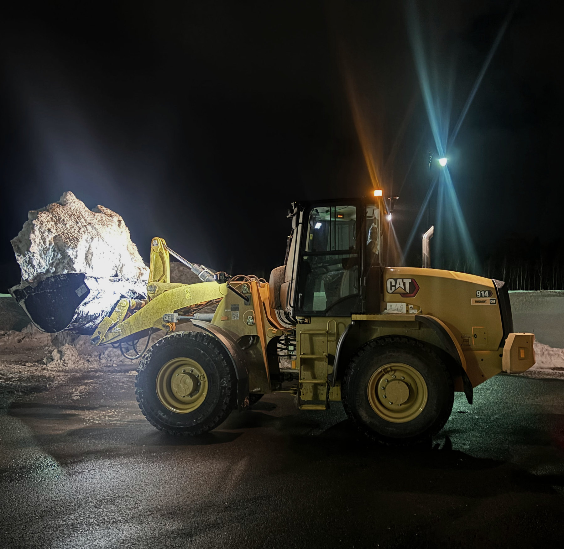 A wheel loader lifting a full bucket of snow at night under work lights, on wet black pavement.