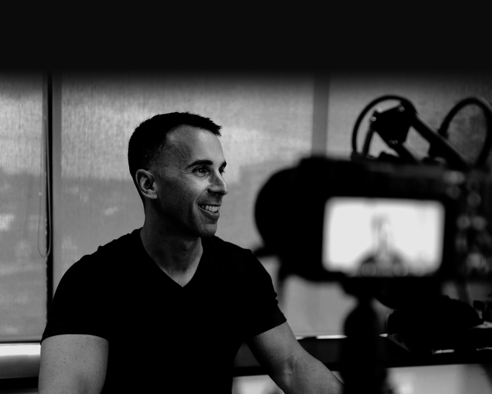 Tony LeBlanc in profile, smiling, being filmed; the camera is out of focus in the foreground, in black and white.