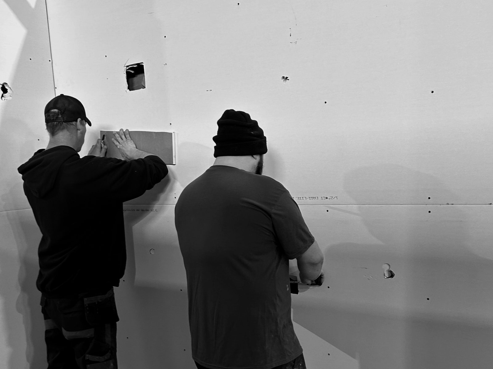 Two of the services company’s workers seen from behind, patching a wall together.