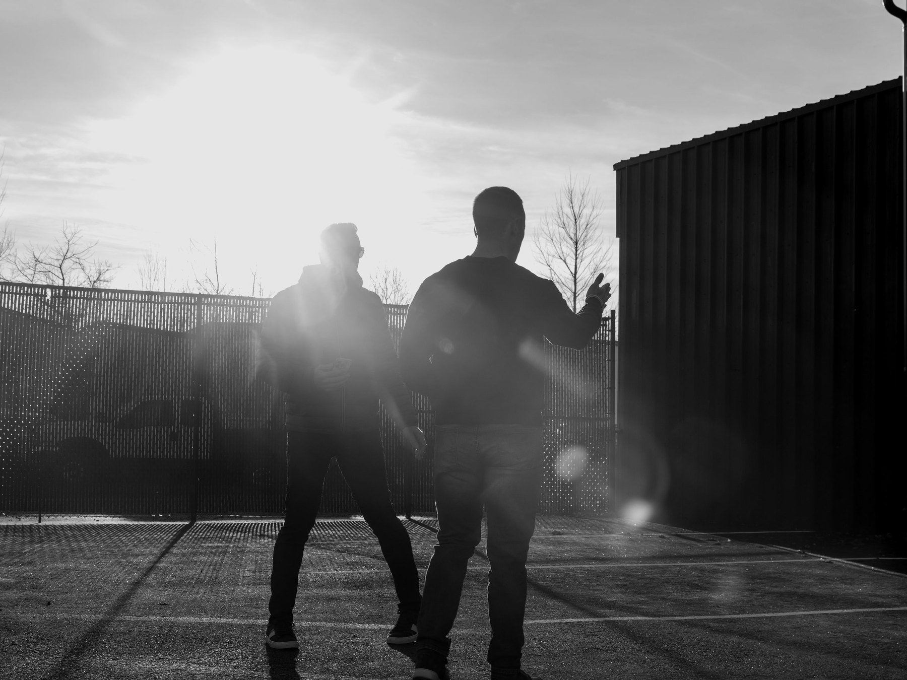 Two people talking as they walk across the services company’s yard into low morning sun, seen from behind, in black and white.