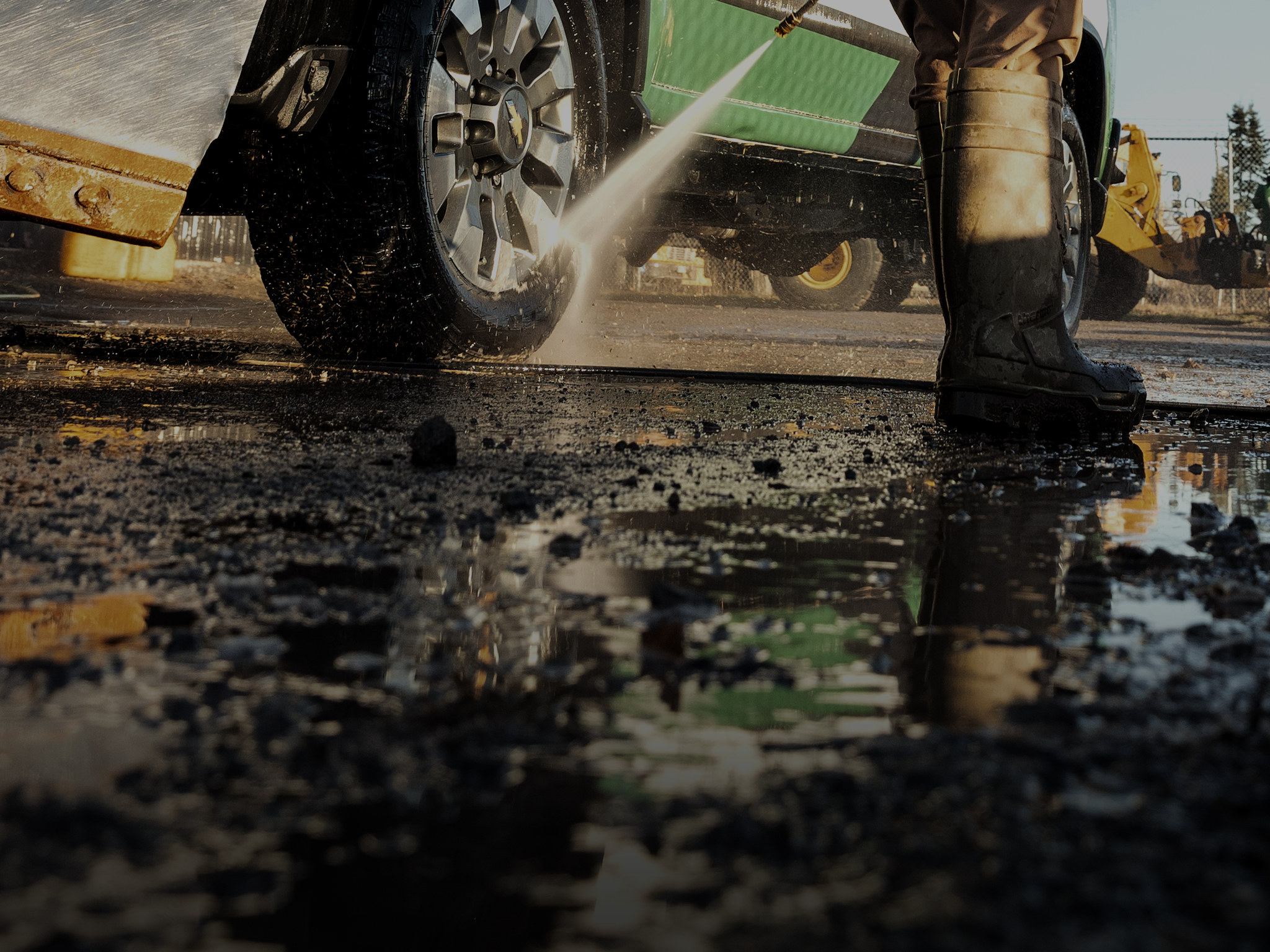 Rubber boots and a pressure washer jet on a truck wheel, water pooling on the gravel.