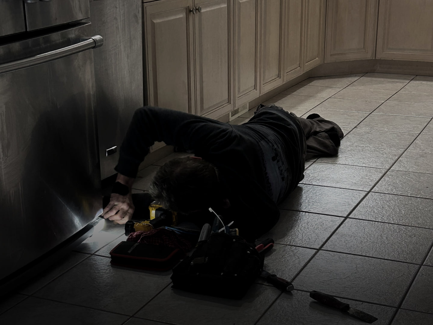 An appliance technician lying on a kitchen floor, working under a fridge by torchlight, his tool bag beside him.