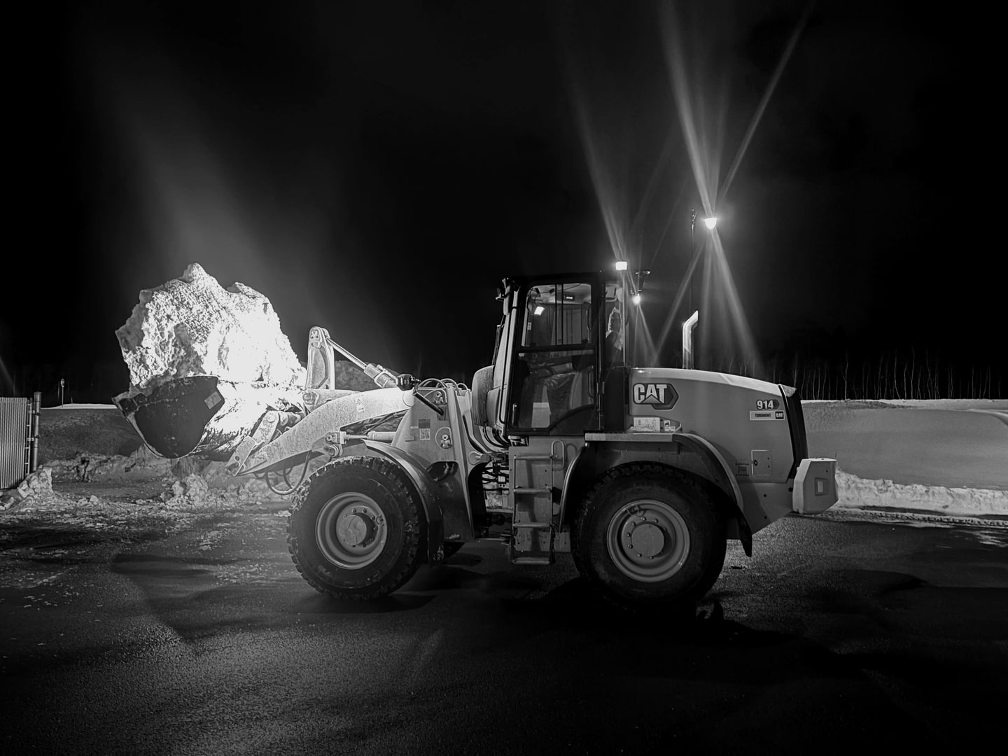 A wheel loader lifting a bucket of snow at night under work lights, in black and white.