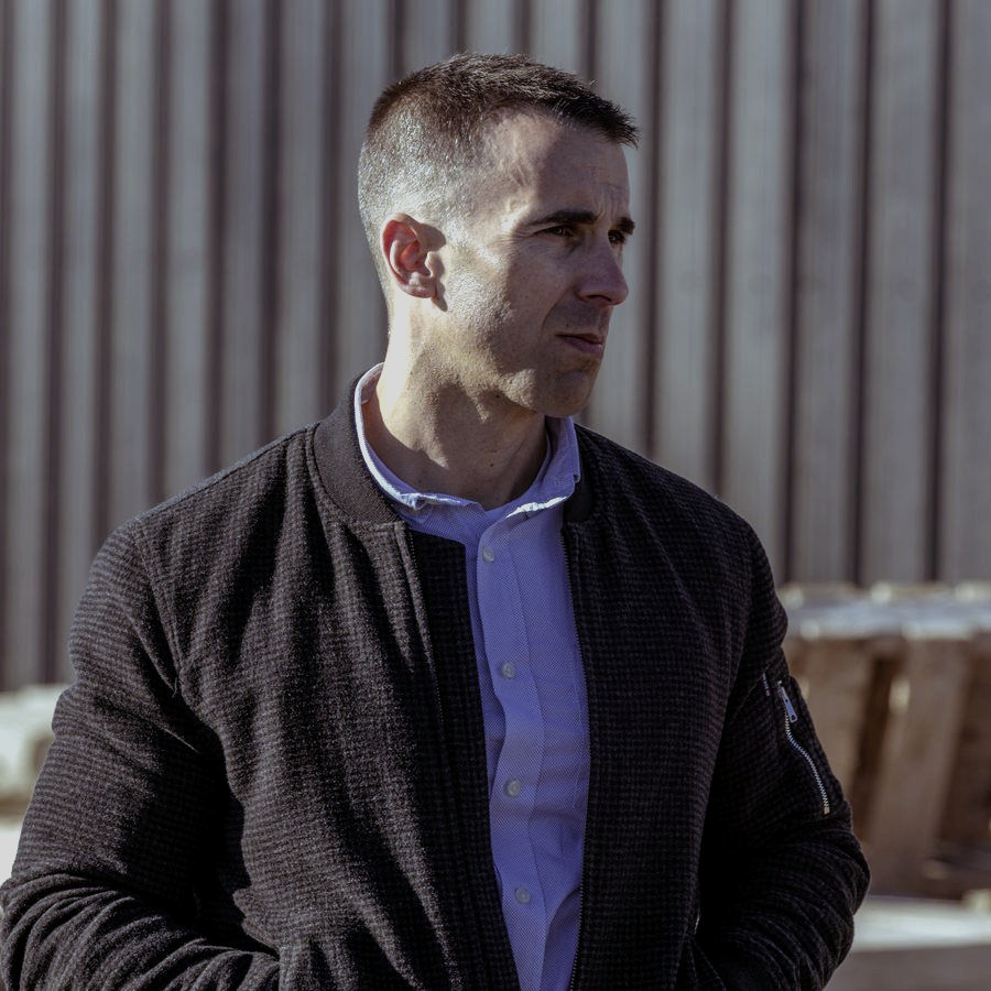 Tony LeBlanc in a dark bomber jacket standing in the company yard in low sun, looking off to one side.