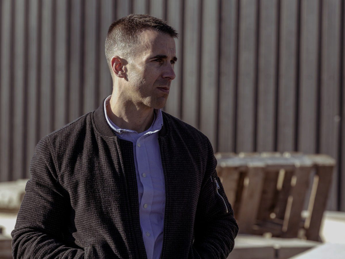 Tony LeBlanc in a dark bomber jacket standing in the company yard in low sun, looking off to one side.