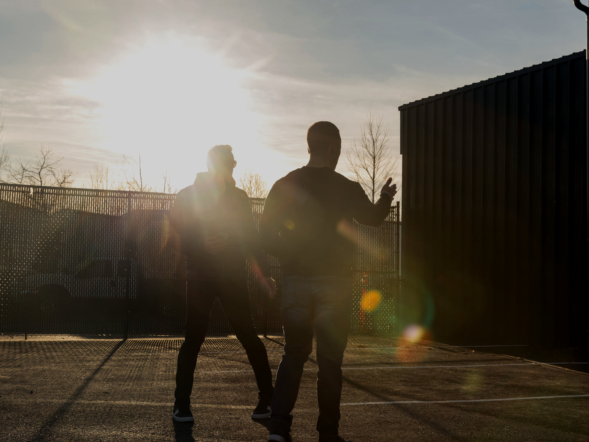 Two people walking across the services company’s yard into low morning sun, a dark steel building beside them.